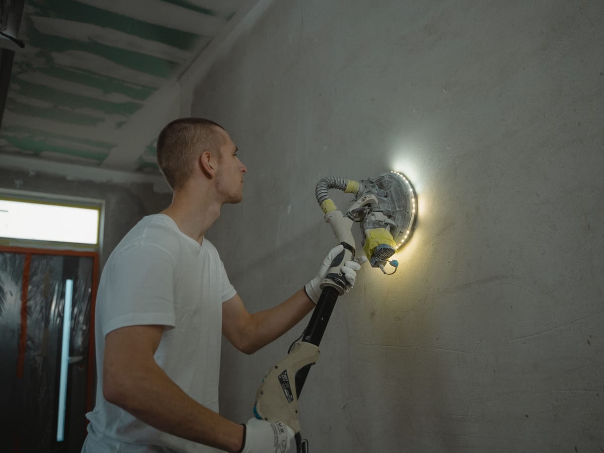 Photo of a man using a power tool to sand a wall during an indoor home renovation project.