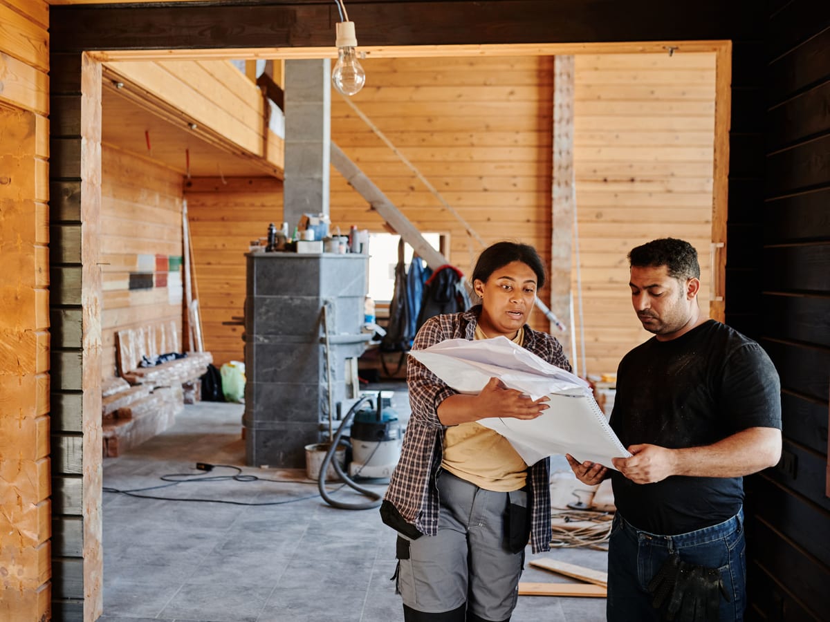 Construction workers reviewing blueprints indoors at a renovation site with wooden walls.